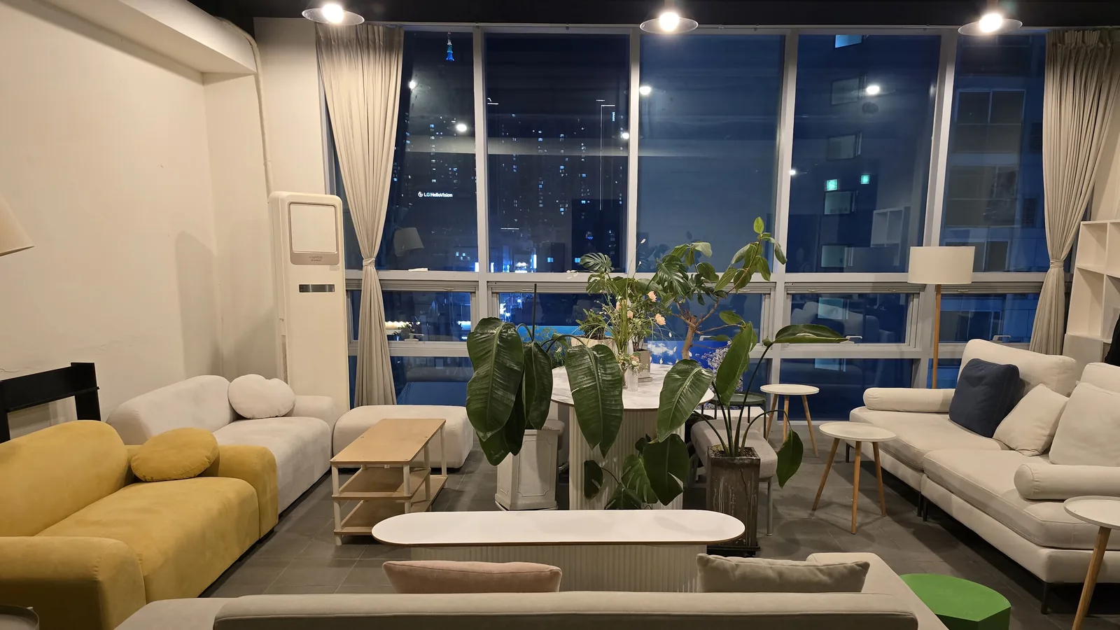 통유리창 앞에 흰 소파와 노란 소파, 타원형 테이블, 큰 화분이 놓인 라운지. 창밖으로 밤 시가지가 보인다